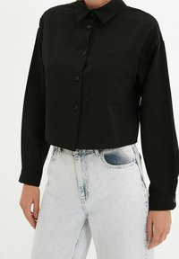 Czarna krótka koszula zapinana na guziki z kieszenią na piersi noszona z wysokim stanem, jasnoniebieskimi dżinsami przez stojącą osobę.