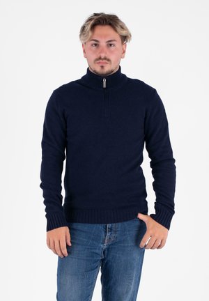 Pull en tricot bleu marine avec un col montant et une fermeture éclair, doté de poignets et d'un ourlet côtelés. Porté avec un jean en denim bleu.