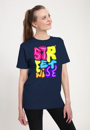 Jeune femme avec queue de cheval brune portant un t-shirt bleu marine avec un texte coloré "streetwise", regardant vers le haut et souriant légèrement.