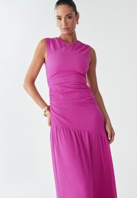 A model poses in a bright pink, sleeveless dress with a fitted bodice and flowy skirt, showcasing a confident expression and stylish accessories.
