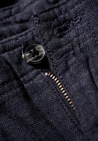 Mörkblått strukturerat tyg med en synlig knapp och metallblixtlås. Tyget har ett subtilt fiskbensmönster.