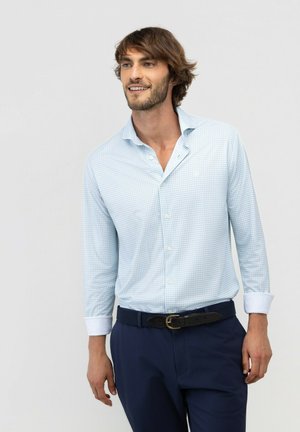 Joven con cabello castaño de longitud media que lleva una camisa a cuadros azul claro y pantalones azul oscuro, sonriendo y mirando ligeramente hacia un lado.