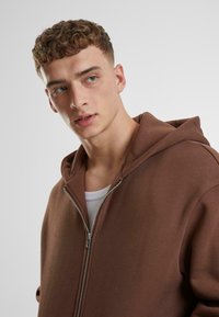 Brauner Reißverschluss-Hoodie aus einem strukturierten Stoff, ausgestattet mit einem Frontreißverschluss, Kapuze und einer darunter sichtbaren Innenschicht.