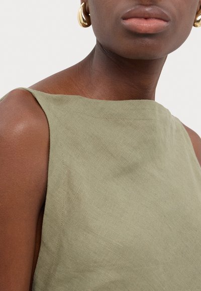 Abito in lino verde oliva con scollatura dritta e spalline larghe, che mette in mostra un tessuto testurizzato e dettagli di cucitura minimi. Orecchini di cerchio dorati.