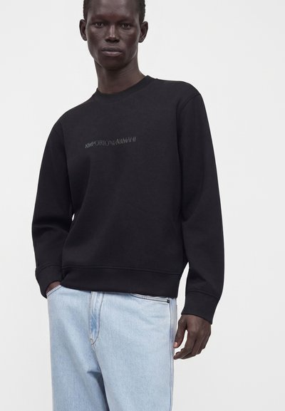 Personne portant un sweat-shirt noir à manches longues Emporio Armani et un jean bleu clair, debout avec une main dans la poche sur un fond uni.
