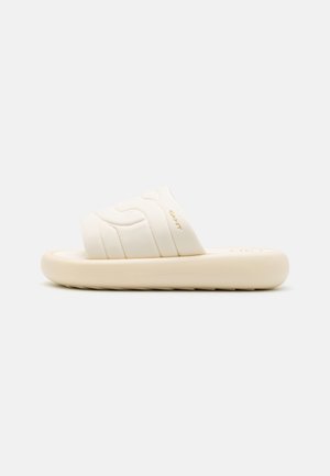 Sandalia tipo slide color crema con parte superior de tela acolchada con diseño acolchado, suela de espuma moldeada y sutil marca en relieve.