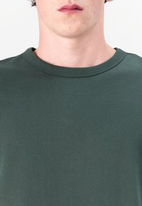 T-shirt en coton vert avec un col rond et des manches courtes. Texture lisse, design ajusté et style minimaliste. Pas de motifs ni d'accents visibles.