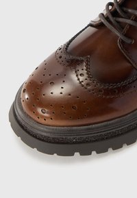 Gros plan d'une chaussure brogue en cuir marron poli avec des perforations décoratives, une semelle noire épaisse et des lacets marron sur fond blanc.