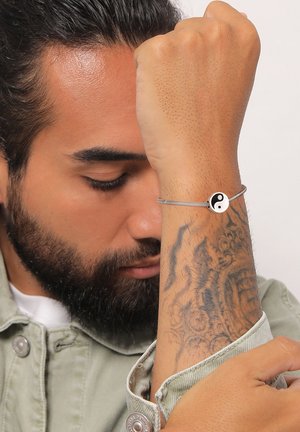 Mand med lukkede øjne, som viser en hævet underarm med et yin-yang armbånd og en detaljeret tatovering, iført en lys denimjakke.