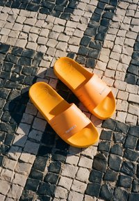 Paire de sandales à enfiler orange vif sur un sol pavé à damier noir et blanc au soleil.