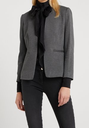 Kvinde iført en tætsiddende grå blazer over en sort bluse med en stor sløjfe, kombineret med sorte bukser, stående mod en ensfarvet baggrund.