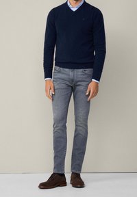 Homme portant un pull bleu marine par-dessus une chemise bleu clair, un jean gris slim et des chaussures en cuir marron foncé à lacets, debout contre un mur uni.