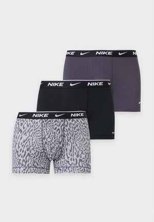 Мъжки модел стои бос със скръстени ръце, облечен в черни боксерки Nike на семпъл светъл фон.
