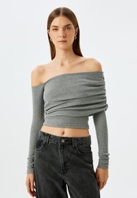 Grauer schulterfreier Langarmtop mit gerafftem Stoff, gerippter Textur und cropped Design, kombiniert mit dunklen Denim-Jeans.