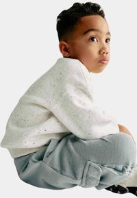 Lichtgrijze gestippelde sweatshirt en zachte blauwe joggers. Het kind is in een hurkende positie, waardoor de structuur van de broek en de naden goed zichtbaar zijn.