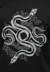 Λευκό αφηρημένο σχέδιο φιδιού που επικαλύπτει παραμορφωμένο γοτθικό κείμενο σε μαύρο ύφασμα, δημιουργώντας μια τολμηρή, γραφική αντίθεση και δυναμικό οπτικό μοτίβο.