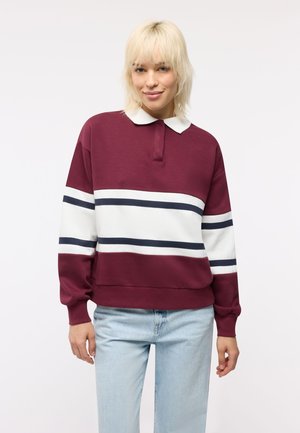 Persona bionda che indossa un maglione a righe bordeaux e bianco con colletto bianco e jeans azzurri chiari, in piedi contro uno sfondo semplice.