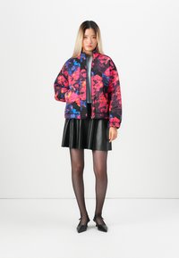 Woman standing wearing a colorful floral quilted jacket, black pleated skirt, sheer black tights, and black pointed-toe heels on white background.