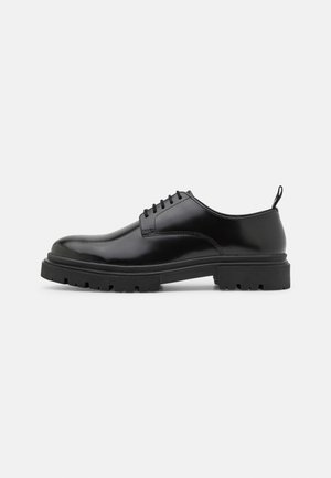 Chaussure noire en cuir à lacets avec un bout arrondi, une finition lisse et une semelle en caoutchouc noir robuste dotée de profondes rainures.