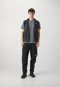 Joven de pie, vestido con un chaleco negro, camiseta gris, pantalones cargo negros y zapatos de senderismo negros, frente a un fondo blanco liso.