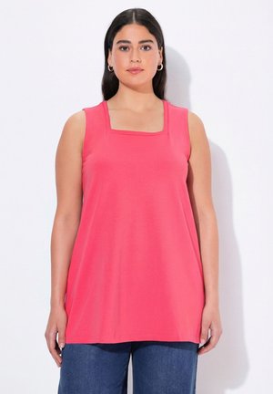 Vrouw met lang donker haar, gekleed in een mouwloze koraalroze top en blauwe spijkerbroek, staand tegen een witte achtergrond.