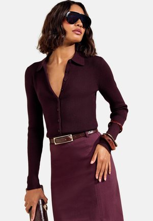 Kvinde iført mørkelilla knaplukket ribstrikket sweater, matchende nederdel, brun bælte, store solbriller og holder en brun tekstureret clutch taske.