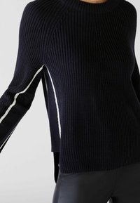 Pull en maille bleu marine avec texture côtelée, présentant des rayures blanches sur les côtés et des épaules tombantes. Le design comprend des fentes sur les côtés pour un détail supplémentaire.