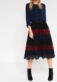 Blusa azul marino con botones combinada con una falda de encaje que presenta franjas horizontales en negro, rojo y azul alternas. Botines negros.