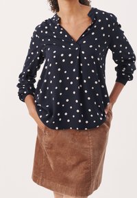 Blusa blu navy a pois con scollo a V e maniche lunghe, abbinata a una gonna in corduroy marrone. La blusa presenta una vestibilità comoda e una consistenza del tessuto morbida.