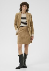 Hnedý kordurový blejzer a sukňa s pruhovaným sveterom pod ním. Čierne kožené čižmy po kolená dokončujú outfit. Celková hladká textúra.