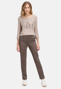 Gestreiftes beigefarbenes Langarmoberteil mit metallischer Beschriftung und hellbraunen Slim-Fit-Hosen aus strukturiertem Material. Das Model trägt weiße Sneaker.
