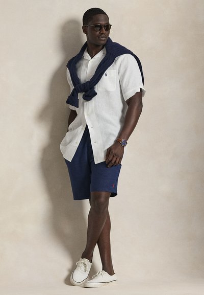 Balta lina īsroku kreklā, tumši zili šorti un gaišas kedas. Tumši zils adīts džemperis pār pleciem; saulesbrilles un pulkstenis kā aksesuāri.