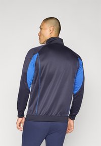 Marineblå og blå sporty jakke laget av glatt stoff. Har høy krage, lange ermer og kontrasterende sidepaneler med blå detaljer.