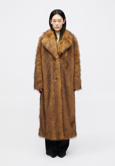 Femme se tenant de face, portant un long manteau en fausse fourrure marron, un pantalon noir et des chaussures noires à bout pointu devant un fond uni.