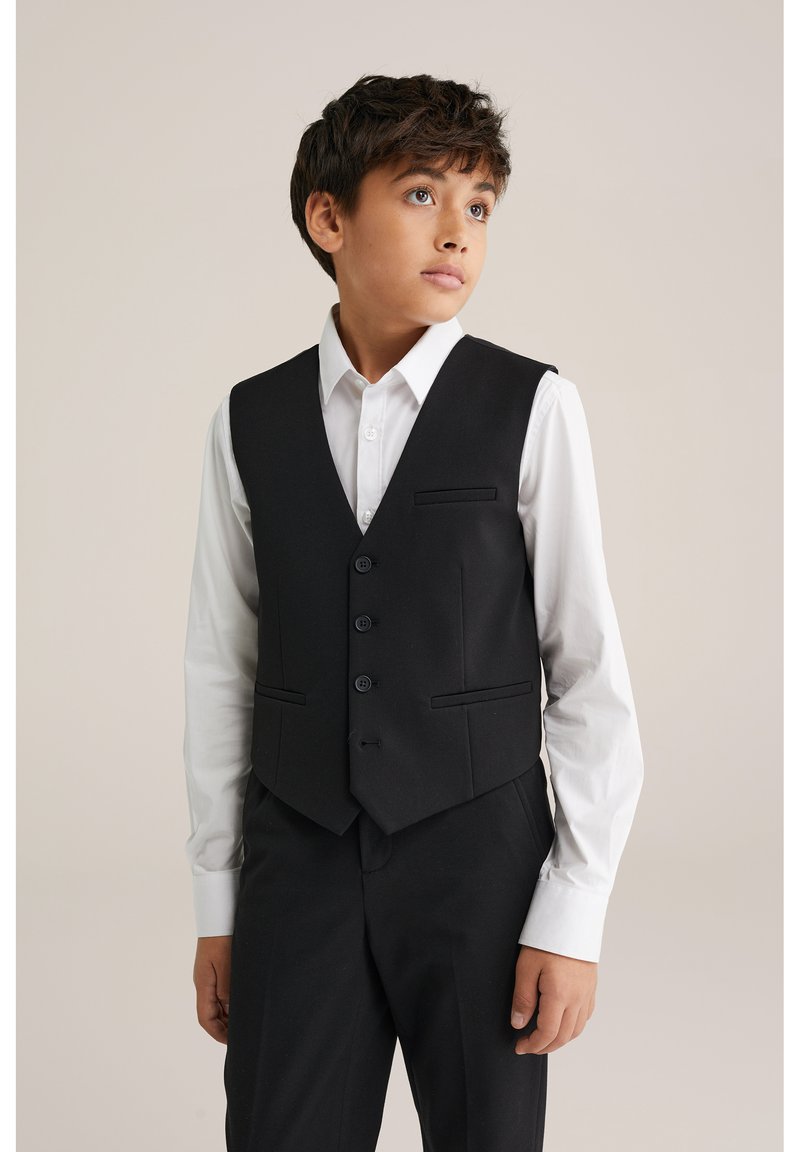 Černá vesta se třemi knoflíky, dvěma předními kapsami a klopou s výřezem, nošená přes bílou košili; střih na míru, hladká textura.
