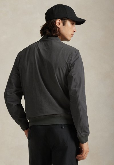 Mees seljaga, kandes tumedat halli bomberjopet, musti pükse ja musta pesapalli nokamütsi tavalise beeži taustaga.