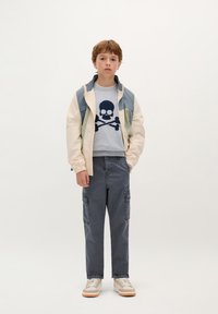 Kurtka bombowca w wielu kolorach, szary sweatshirt z czaszką, szare spodnie cargo, beżowe sneakersy z pomarańczowymi akcentami, jednokolorowe tło.