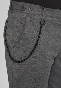 Pantalon gris texturé avec une chaîne noire attachée à la passoire de ceinture et à la poche, porté avec une chemise noire visible à la taille.