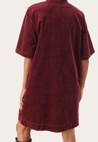 Übergroßes Kleid aus burgunderrotem Cord mit kurzen Ärmeln, geradem Schnitt und einer strukturierten Oberfläche. Der Saum endet mit einer dezenten Umschlagkante.