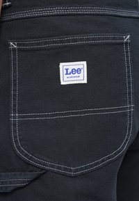 I pantaloni da lavoro in denim neri presentano una tasca posteriore distintiva con un'etichetta bianca che recita "Lee Workwear". Le cuciture a contrasto evidenziano i bordi delle tasche.