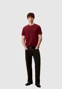 Burgunderrotes Baumwoll-T-Shirt mit Rundhalsausschnitt, kombiniert mit schwarzer Hose und schwarzem Gürtel. Das Modell steht mit den Händen in den Taschen und trägt schwarze Schuhe.