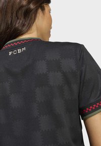Donna che indossa una maglia sportiva nera con un sottile motivo a croce, colletto e bordi delle maniche a quadri rossi e oliva, e il testo "FCBM" vicino al collo.