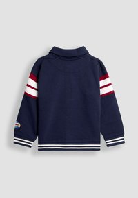 Námornícky modré mikina s golierom, bielymi a červenými pruhmi na rukávoch a pruhovaných manžetách. Obsahuje logo nášivku na rukáve.
