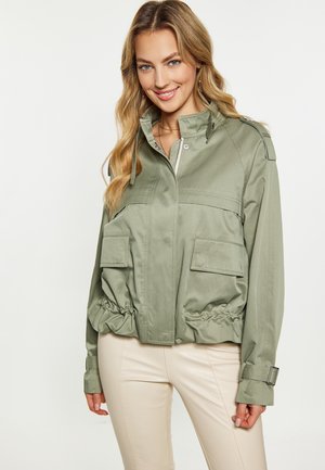 Woman wearing light green utility jacket with front pockets and beige pants, smiling with long wavy blonde hair against plain background.