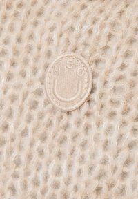 Beige gestrickter Stoff mit einer lockeren Textur, ausgestattet mit einem weichen, runden Logo-Patch, das in der Mitte die Buchstaben "HGO" eingeprägt hat.