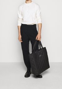 Svart kviltad tote-väska med en minimal design, med en logotypdetalj. Modell bär en vit skjorta med färgglada fållar och svarta byxor.