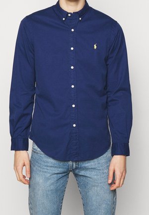 Homme portant une chemise bleu marine à manches longues boutonnée avec un petit logo jaune sur la poitrine et un jean bleu clair, debout.