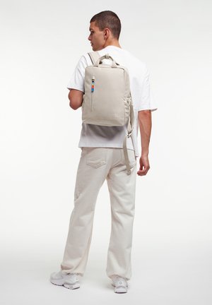 Mies, jolla on valkoinen t-paita, vaaleanbeiget housut ja valkoiset lenkkarit, kantamassa vaaleanbeigettä suorakulmaista reppua, jossa on värikäs vetoketjunnaru.