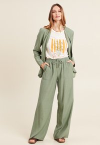 Blazer vert clair sur un t-shirt blanc avec un graphique orange et vert, associé à un pantalon large ample assorti et des sandales marron.