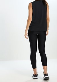 Žena v bezrukávovém černém topu, černých zkrácených legínách a černých sportovních botách stojí s otočením zády k fotoaparátu.